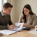 Un jeune homme et une jeune femme étudient ensemble un dossier de financement immobilier avec des graphiques abstraits et une calculatrice sur un bureau sobre.