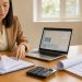 Investisseuse préparant un dossier de financement immobilier sur un bureau lumineux avec des documents vierges, un ordinateur portable et des plans architecturaux.